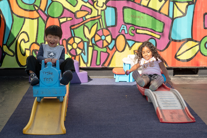 A child on the left rides a little blue train down a plastic yellow slide. The child on the left slides down a red and white plastic slide.