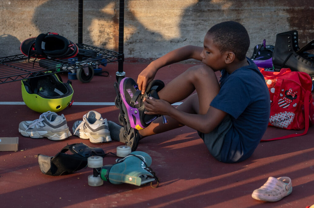 A boy straps on a pair of purple Rollerblades, stray sneakers and skates littering the ground around him.