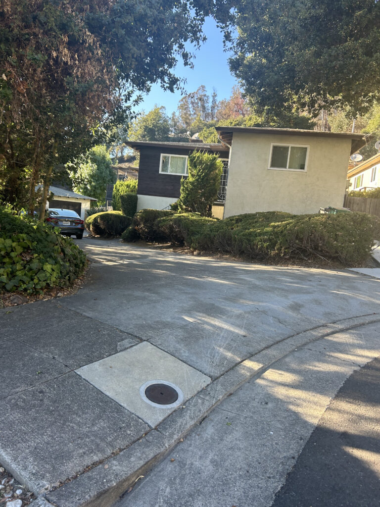 A brown and beige two-story home sits at the top of a driveway on a curved street, a canopy of trees overhead.