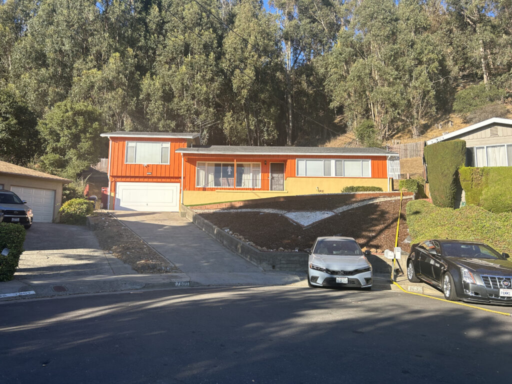 And orange and white long one-story house sits atop a low hill on a curved street, with towering green trees behind it.