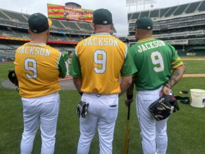 Three baseball players with their backs to the camera look at the field in front of them. Two are wearing gold shorts and one green. They all say Jackson 9 on the back