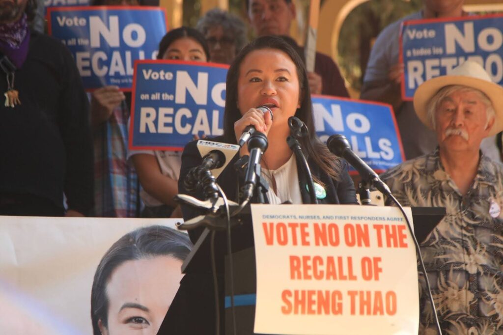 Mayor Sheng Thao speaks into microphones at a ralley, with people holding"vote No on the Recall" signs behind her.