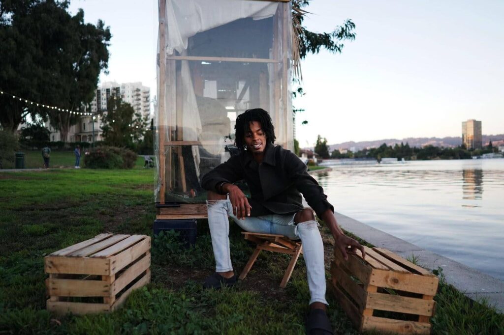 A man sits on a wooden chair with two wooden crates on either side of him beside a lake on which stands his makeshift wooden and plexiglass cabin.