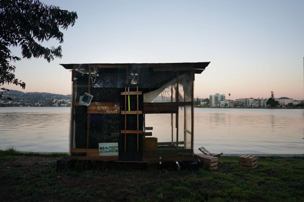 A makeshift cabin sits on the banks of Lake Merritt, with the lake and Oakland hills behind it.