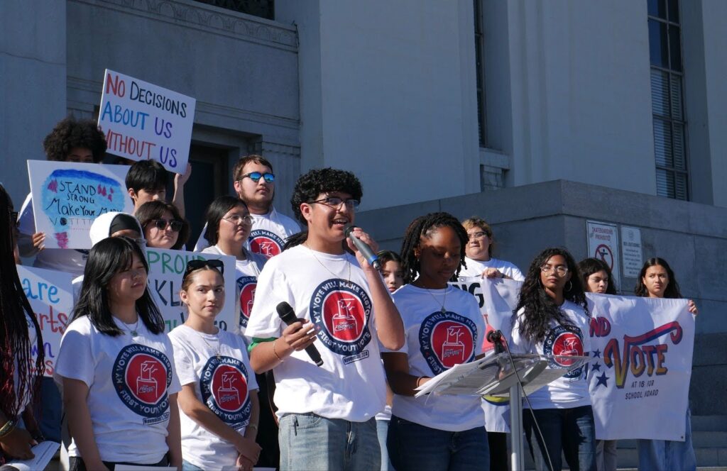 A group of teens in white T-shirts with a red and blue ballot logo on them hold signs and stand outside a palatial building. A boy and a girl in the center are standing at microphones.