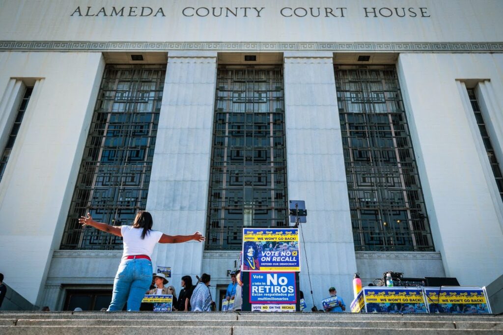 A woman stand with her back to the camera, arms outstretched facing a crowd in front of a white-pillared building.