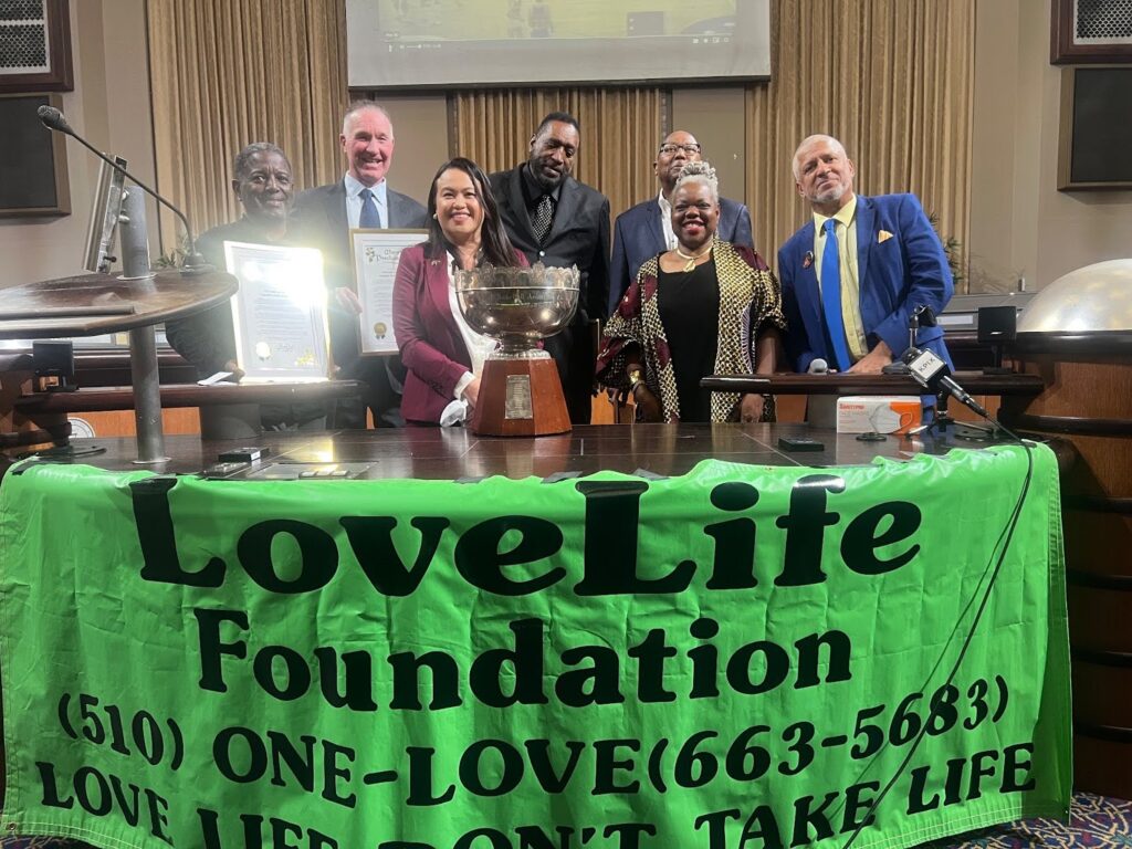 Seven people stand behind a table with a green banner in the front that reads Love Life Foundation.