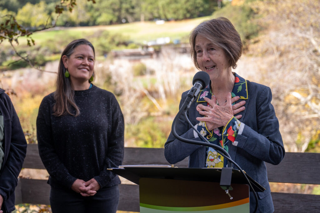 A woman stands at a podium with both hands on her chest, mouth open in mid-sentence in an outdoor wooded setting. Another woman on her left is smiling at her.