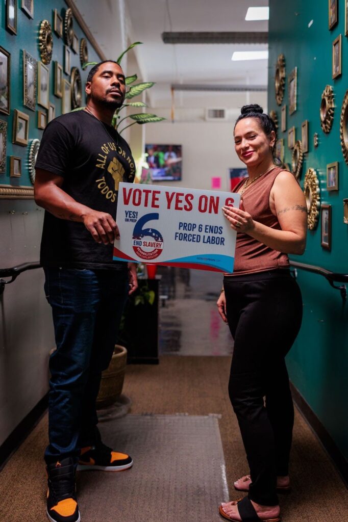 Two people stand in a hallway, a man on the left, a woman on the right, holding a vote yes for Prop 6 sign.