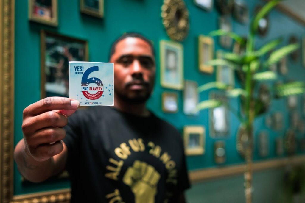 A man is blurred in the background but in his outstretched right hand is a clear patch that says yes on prop 6