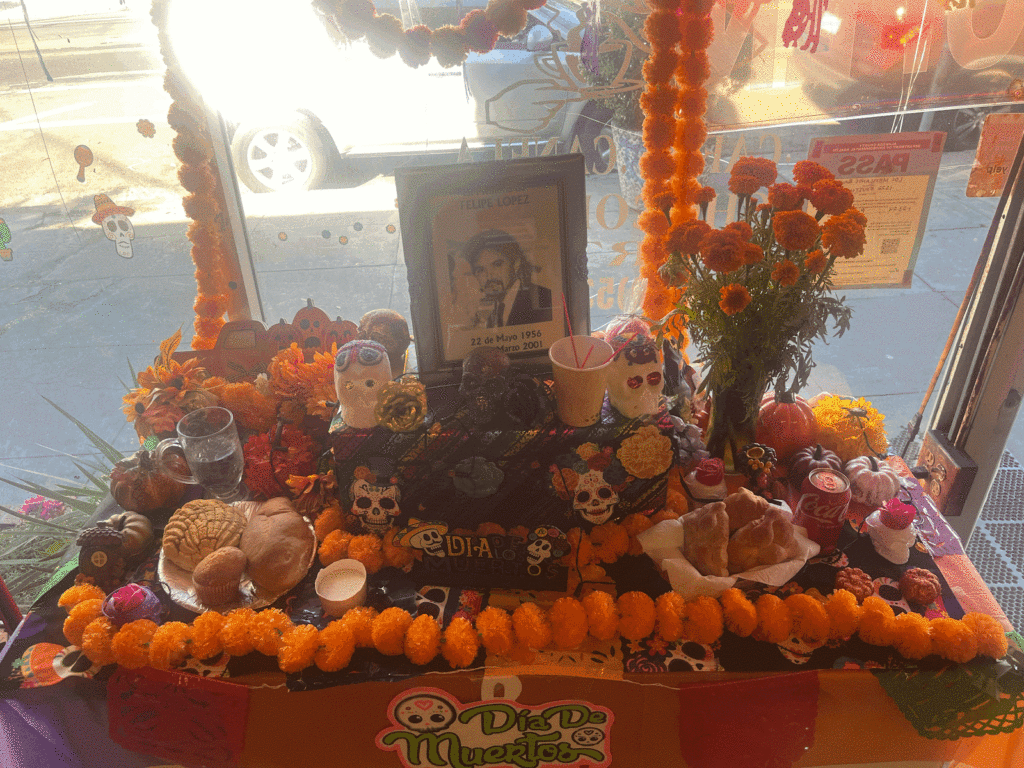 An altar in a storefront window includes marigolds around the edges and in a vase, candles, skulls, bread, a can of Coke, a glass of water and a photo of a man with a mustache.