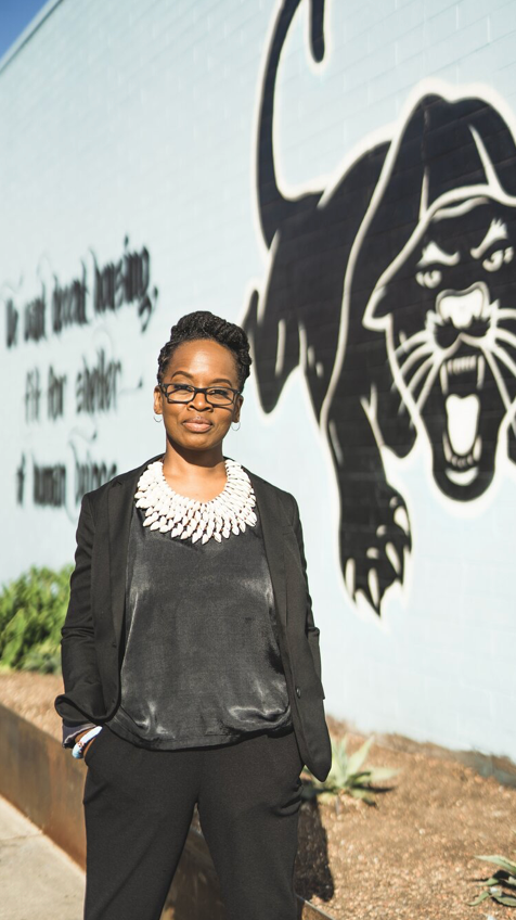 A woman in black except for a white beaded collar stand in front of a lion mural