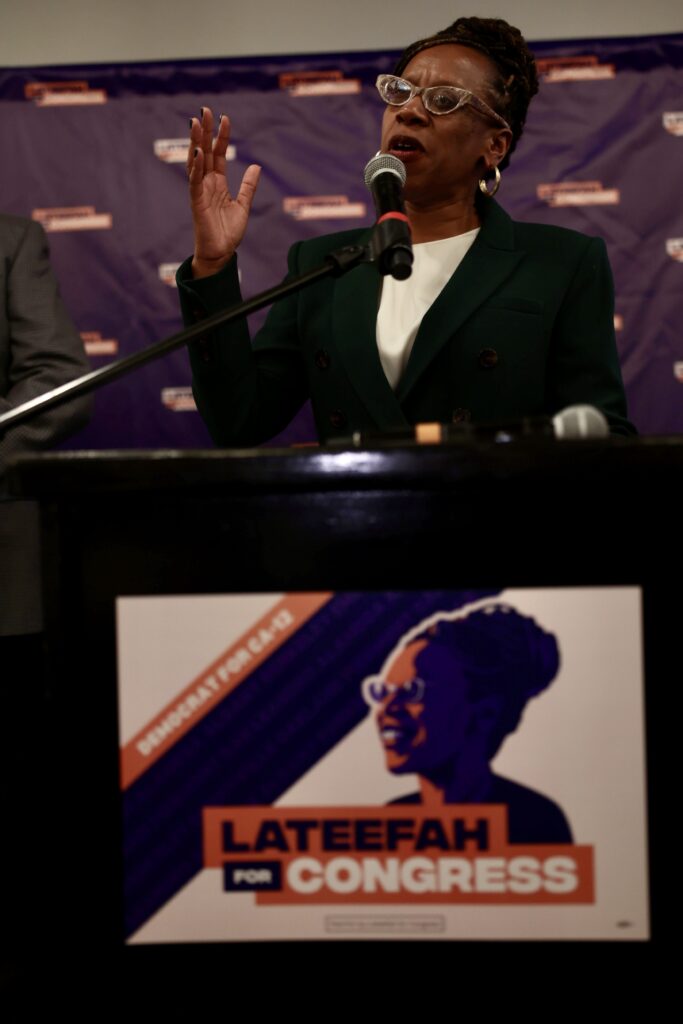 A woman stands at a microphone behind a podium and talks with her right hand raised.