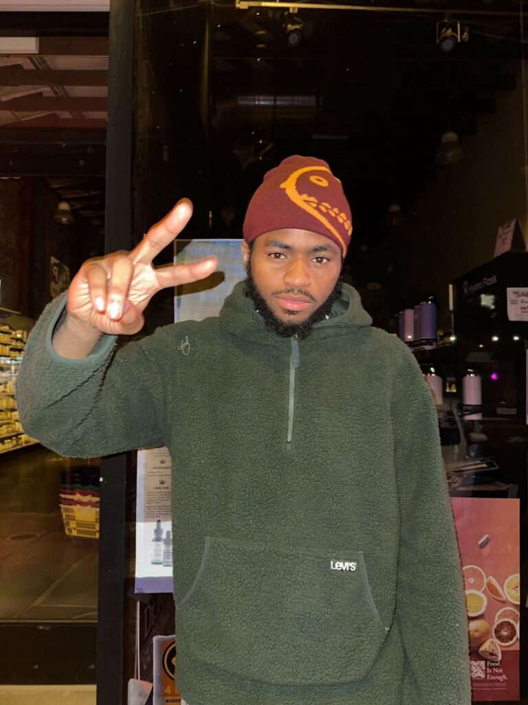 A man in a fuzzy hunter green hoodie and an orange knit hat is looking at the camera, flashing a sideways peace sign with his right hand.