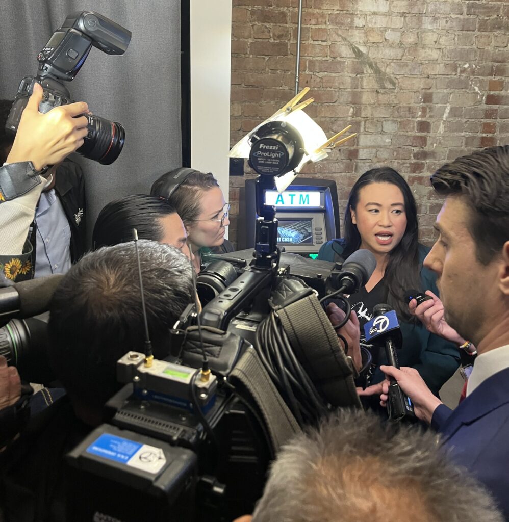 A woman is caught in mid sentence with her mouth open, surrounded by a throng of reporters with camera and microphones in a small, brick-walled room.