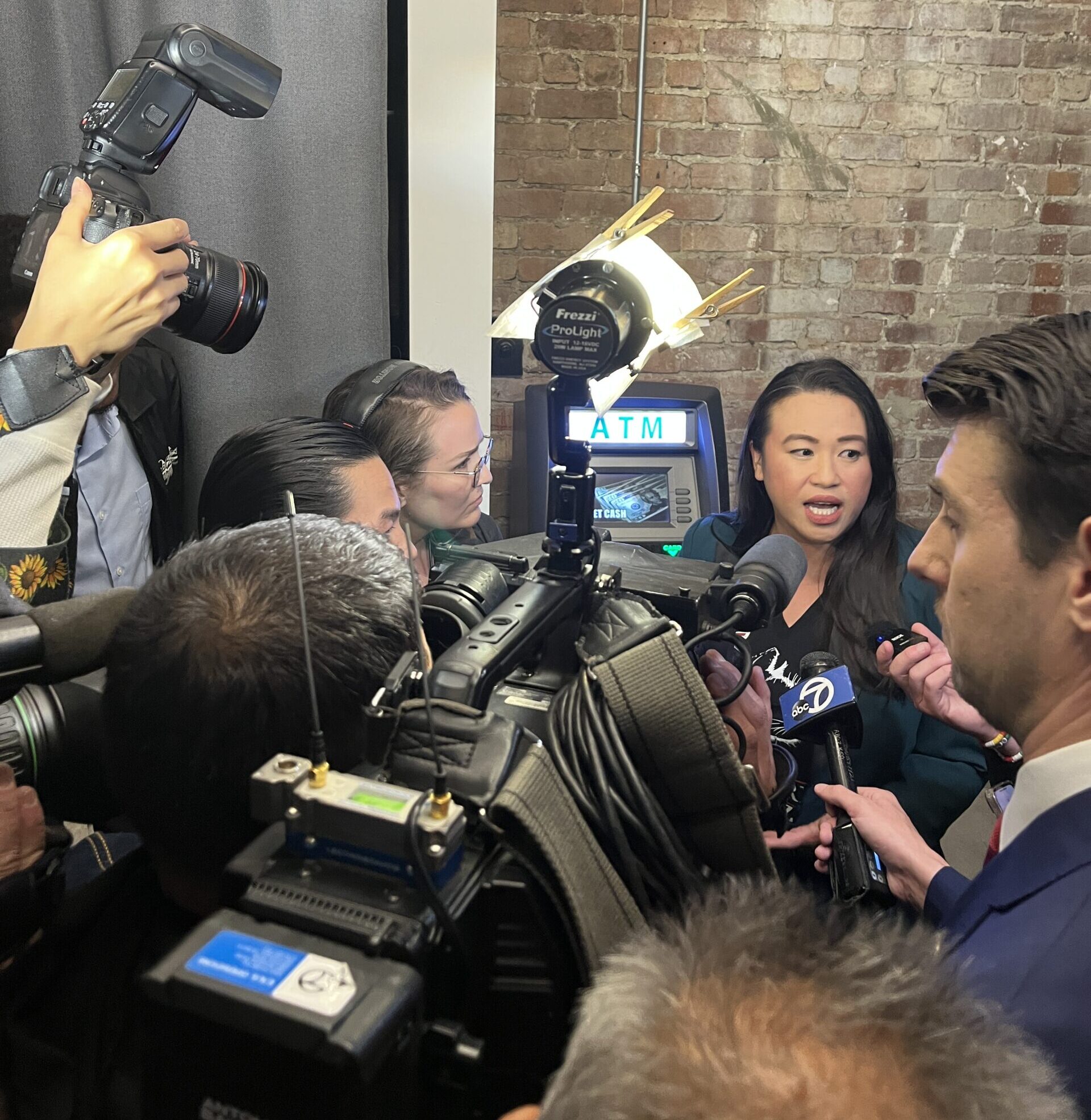 A woman is caught in mid sentence with her mouth open, surrounded by a throng of reporters with camera and microphones in a small, brick-walled room.