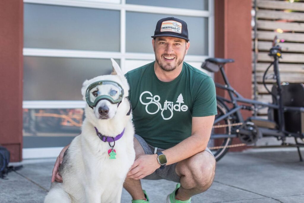 A man with a green T-shirt and cap crouches next to a white dog wearing a green visor.
