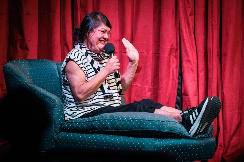 A woman dressed all in black and white sits with her feet up on a couch on a stage, holding a microphone.
