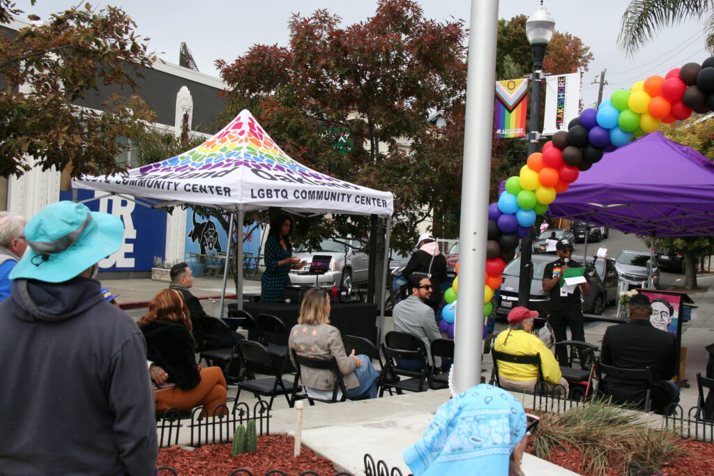 Under an arch of rainbow balloons and a purple canopy stands a man dressed in black except for a rainbow badge, with a microphone reading something to an audience of about a dozen people in folding chairs on a city sidewalk.