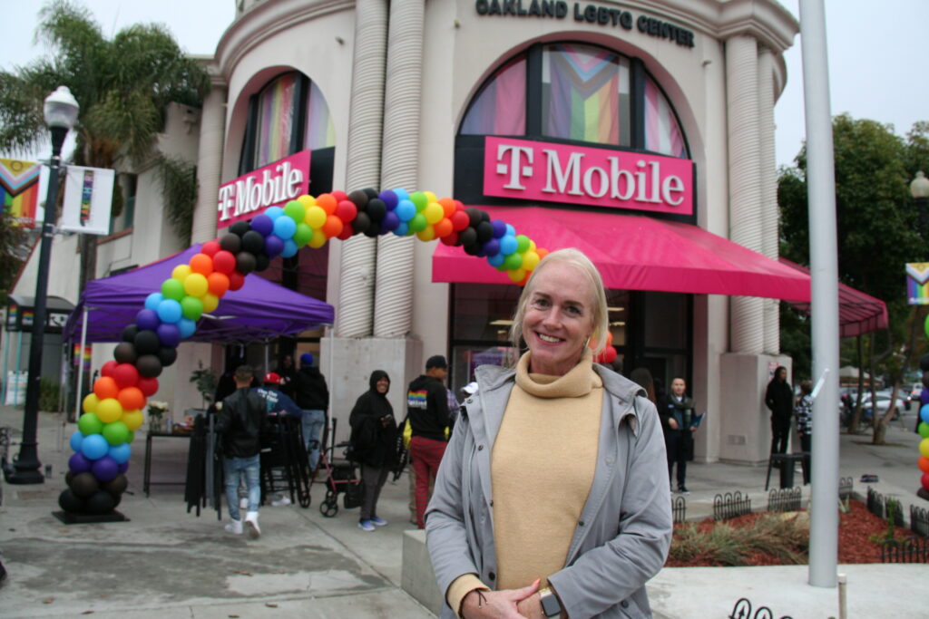 A woman with blond hair and a tan turtle neck sweater smiles outside a T-Mobile store where an arch of rainbow balloons is set up.