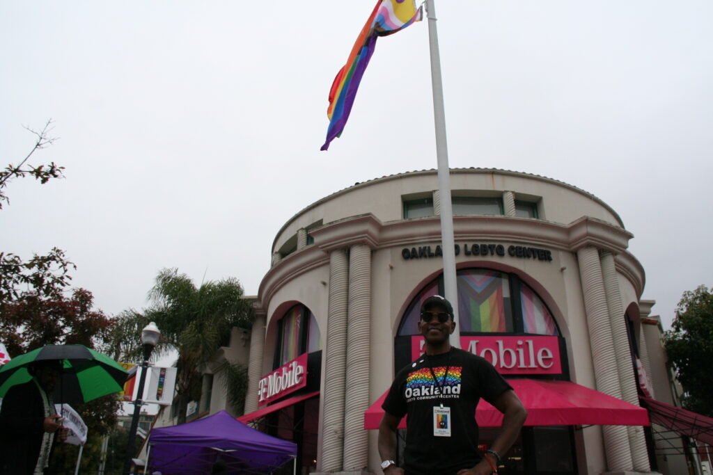 A man wearing a black cap and black T-shirt with a rainbow on it stands with his hands on his hips under a rainbow flag outside a T Mobile store.
