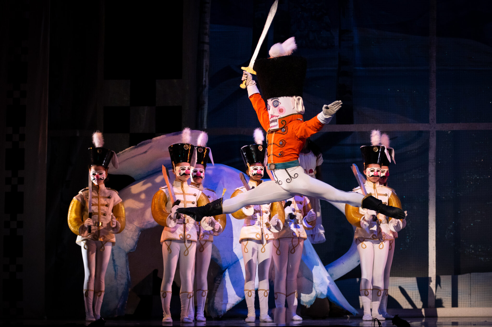 On a stage, four lines of toy-soldier-like characters are standing at attention in mostly white outfits, with yellow and white jackets and tall black hats. In the middle of the frame, a red-jacketed solider with a sword in his right hand is leaping before them, his legs in a mid-air split.