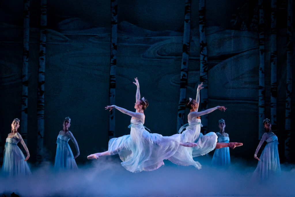 Two ballerinas facing opposite directions, both in flowing white robes are captured in a mid-air leap, their legs outstretched in splits above white smoke, while four other dancers stand behind them in similar gowns on a dark stage.