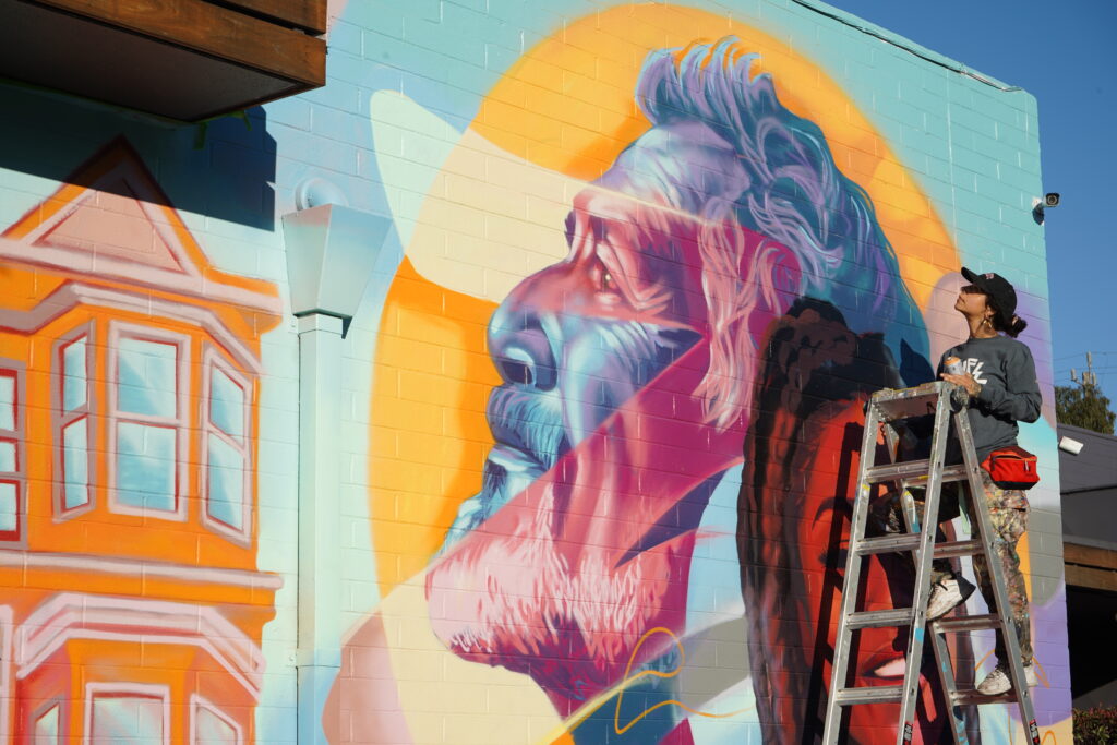 A painting on the side of a brick building depicts that large head of a bearded man with wavy hair in blue with red and yellow streaks, looking left, and a smiling woman with long dark braids looking straight ahead. In front of the wall, a person in a baseball cap holding a can is facing the mural and standing on a ladder.