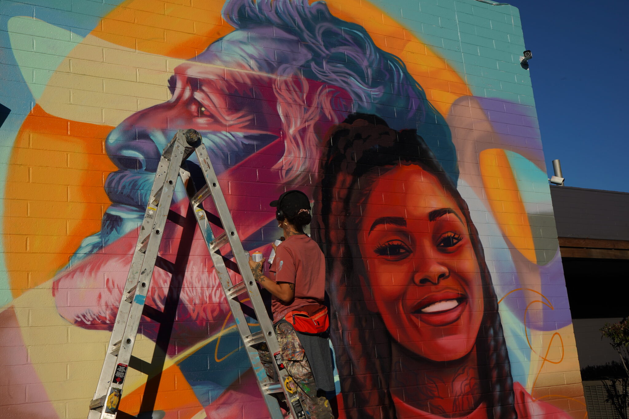 A painting on the side of a brick building depicts that large head of a bearded man with wavy hair in blue with red and yellow streaks, looking left, and a smiling woman with long dark braids looking straight ahead. In front of the wall, a person in a baseball cap holding a can is facing the mural and standing on a ladder.