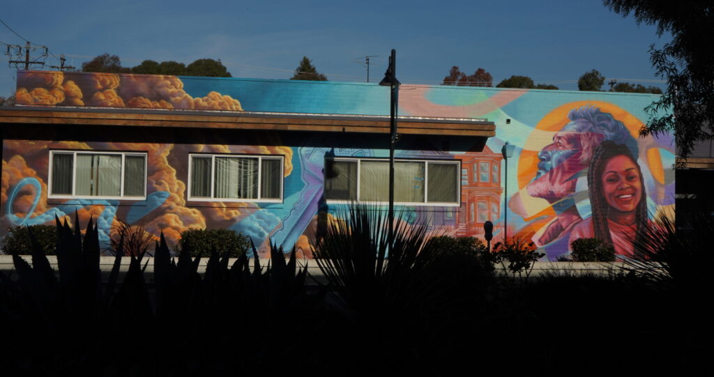 A painting on the side of a brick building depicts that large head of a bearded man with wavy hair in blue with red and yellow streaks, looking left, and a smiling woman with long dark braids looking straight ahead. To their left, a Victorian home is painted in red.