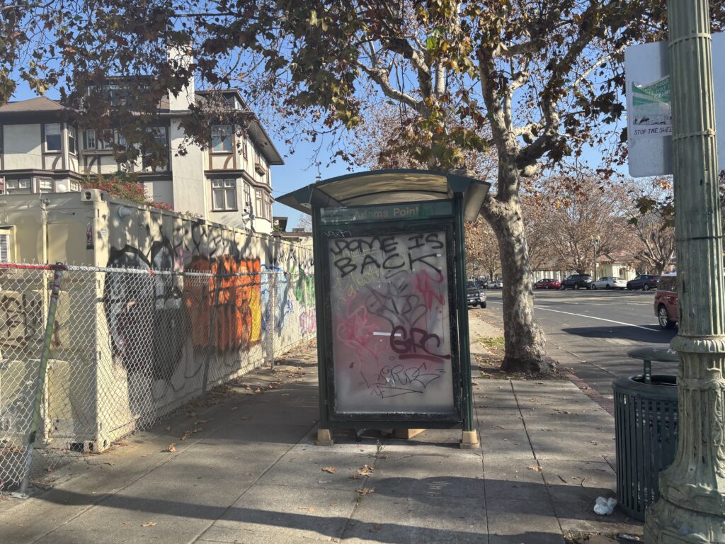 In the middle of the frame is a square bus shelter with graffiti painted on it, on a sidewalk by a tree and a chain-link fence.