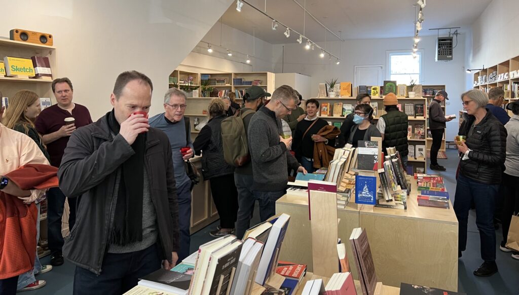 About 20 people are milling about a crowded room full of pale wooden bookshelves and in the front, a man sips from a red cup.