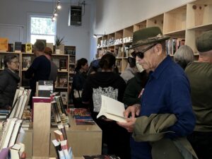 About 20 people are milling about a crowded room full of pale wooden bookshelves and in the front, a man wearing a fedora and sunglasses is reading a book.