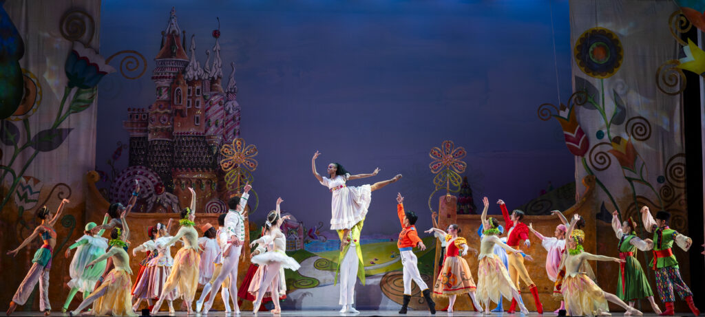 A stage set with a castle on the left and large flowers on the right. At the forefront are dancers in bright colors, about eight on each side of the main dancer, who is at center stage in a white gown, being lifted by another dancer.