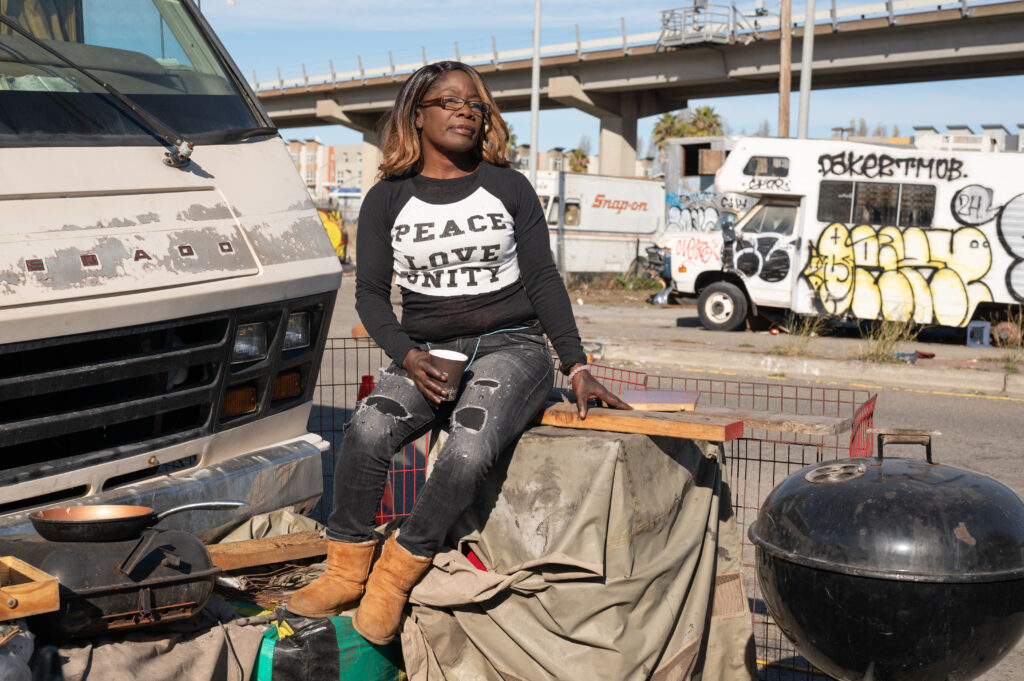 A woman in a black and white shirt and ripped black jeans sits on a tarp covering her possessions, next to an RV in an encampment.