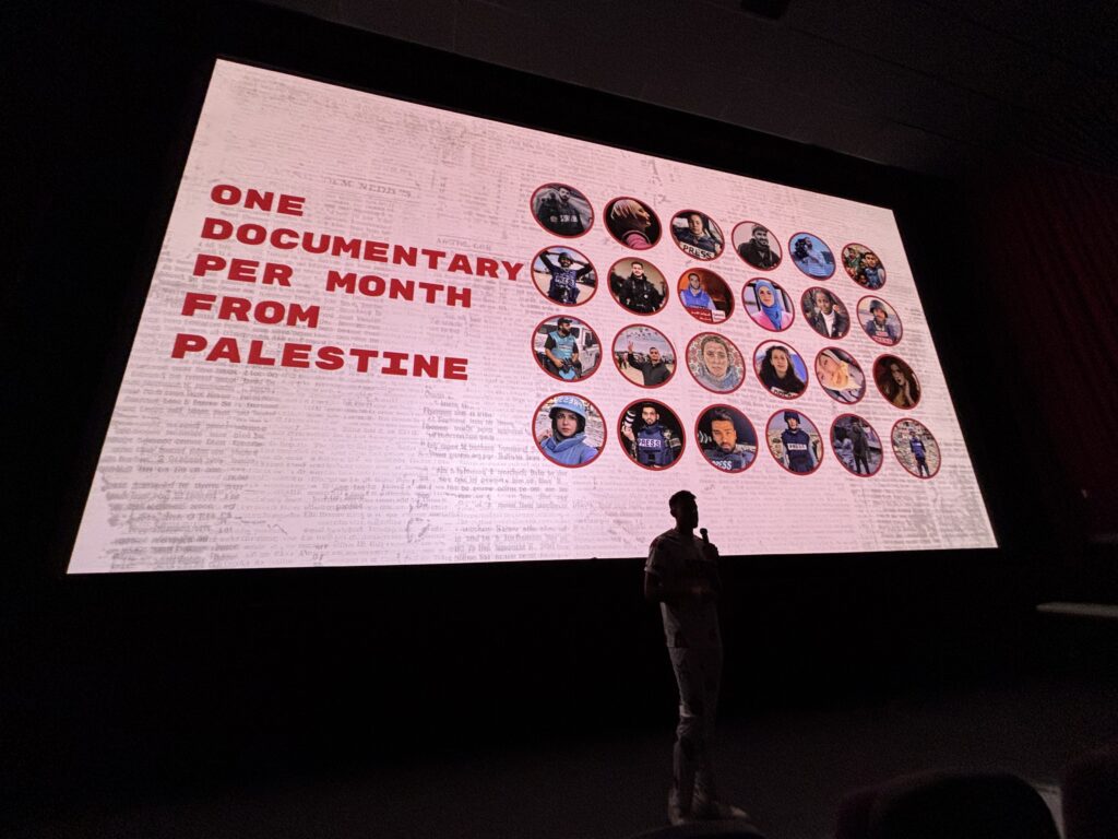 A man with a microphone is silhouetted against a projected image that has some news text and 24 circles stacked 4 x 6 with people's faces in them.