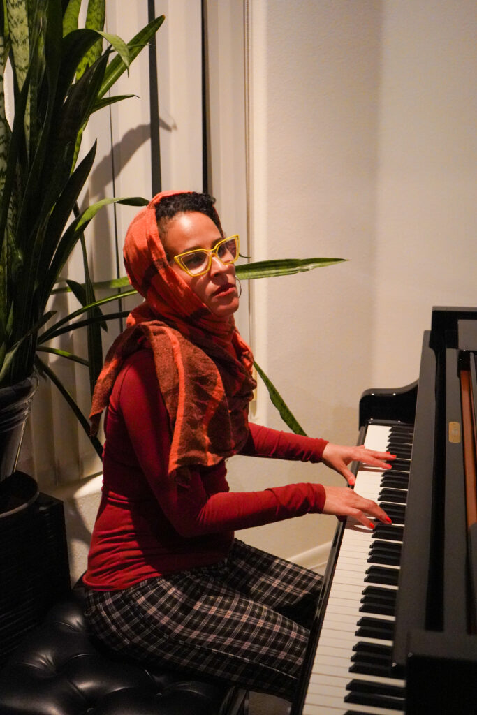 A woman in a head scarf, glasses, and red shirt plays at a piano.