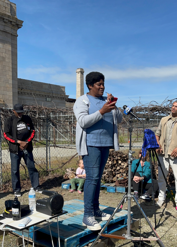 Under a blue sky, a woman in jeans, sneakers and powder blue T-shirt under a gray sweater looks at her phone as the stands before a microphone in front of a chainlink fence and on top off a makeshift square stage made of wooden crates.