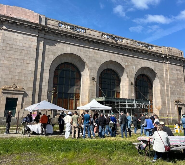 Outside a stately old beige-bricked building with three curved archways stands a few dozen people in front of two tables with white awnings over them. In the foreground, a woman sits at a picnic table on the lawn.
