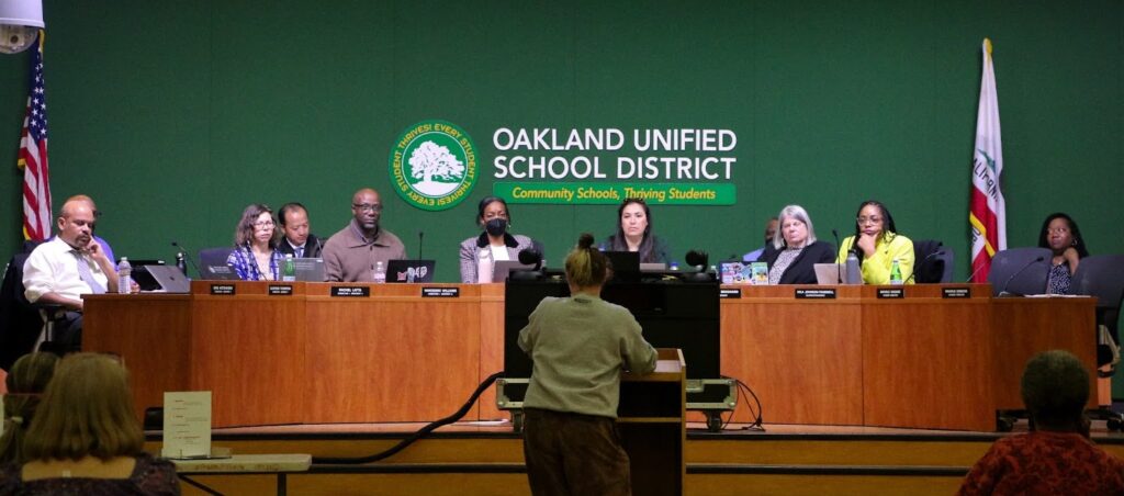 Behind a long brown table sits six members of the Oakland School Board, then the superintendent on the far right, next to an empty chair. Behind them is a sign saying Oakland Unified School District, with a tree logo. On either side of the table are flags on poles, the American on the left, and the California on the right.