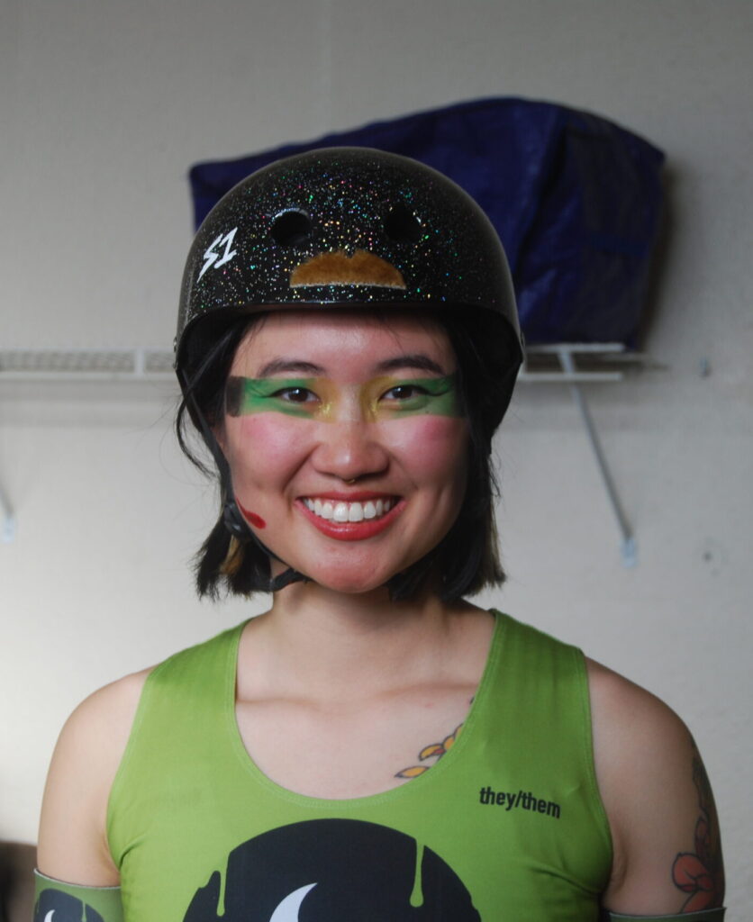 A person with short black hair, a black sparkly helmet, green shirt, and green eye makeup smiles at the camera.