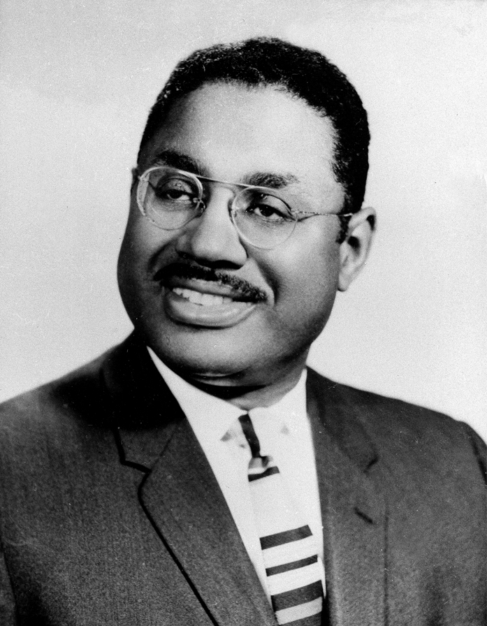 A black and white head and shoulders shot of a man with close cropped dark hair and a mustache, wire-rimmed glasses and smiling, his head tilted toward the left, wearing a horizontally striped tie over a white shirt and under a dark suit jacket.