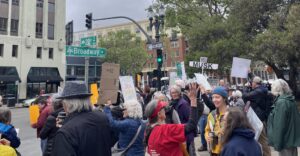 A group of about 50 people, many with signs, stand on a busy street under intersecting signs that say 22nd Street and Broadway