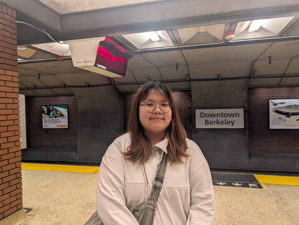 A woman in shoulder-length dark hair with the bottoms sort of blond and wearing glasses stands in a white zip-up jacket, a denim bag slung crosswise over her body, on a subway platform. A brick wall is in the distance and an electronic sign above her head shows red letter.