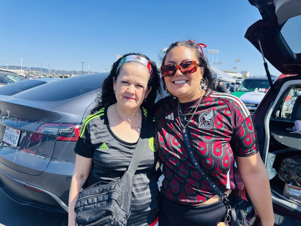 Two women smile for the camera in a parking lot. The one on the left is older and wearing a gray and green adidas T-short, her long curly hair pulled back in a Mexican flag bandana. The younger woman on the right is wearing red and green ribbons in her dark hair, sunglasses and a red and green shirt.