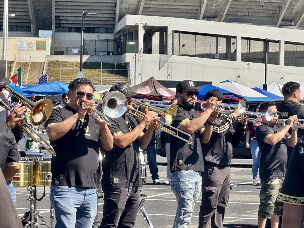 Six horn players in a band ppay trombones and trumpets in a parking lot outside a stadium. All are wearing black T-shirts, jeans and sunglasses.