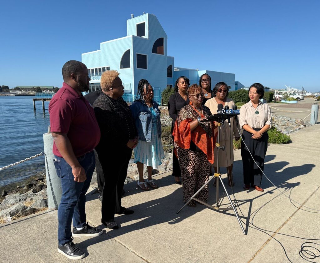 A woman in flowing orange and leopard-patterened dress, short curly hair and glasses stands at a microphone on a sidewalk. Six people are standing on either side of her. Behind them is a blue geometric building on a waterfront.