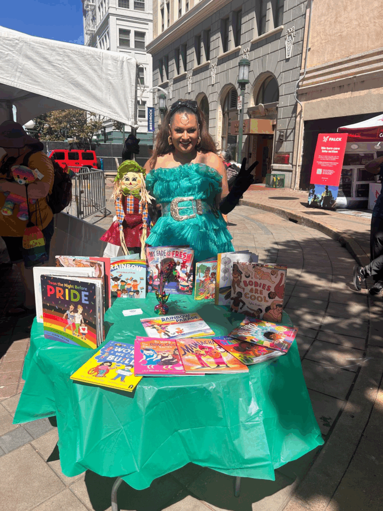 A drag artist in long auburn hair held back by a cloth band wears a turquoise sleeveless outfit with a large gold belt and stands before a table filled with children's books on an Oakland street.
