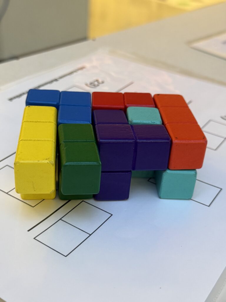 A three-dimensional figure using small yellow, blue, green, red and turquoise squares is pictures atop a white sheet of paper with squares inked in.