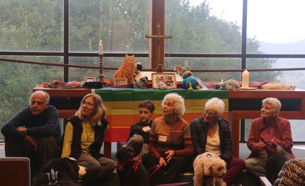 From left, a man, woman, child about 5 years old, followed by three more women sit in folding chairs in front of an altar topped with a Bible, cross and stuffed animals. Three dogs are sitting either on or in front of the three people in the center of the photo.